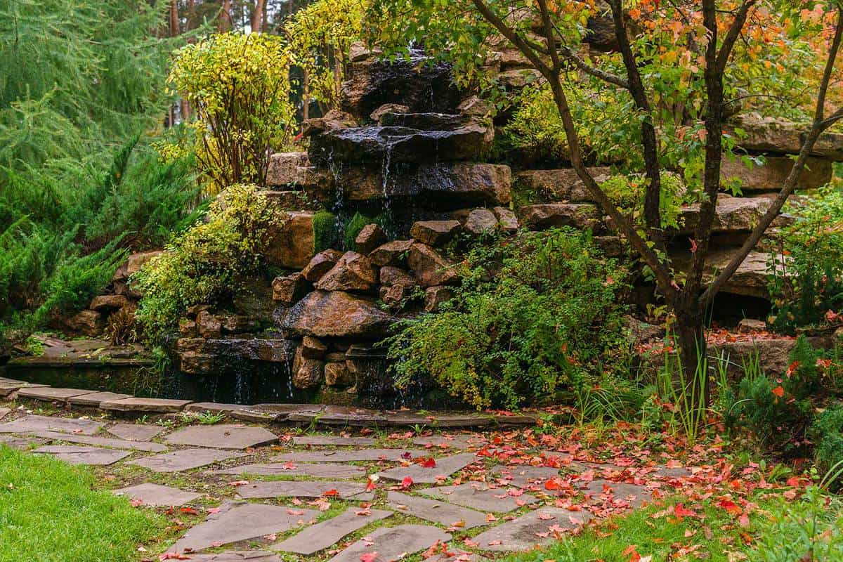 Lawn among decorative bushes with a path and artificial rock with waterfall