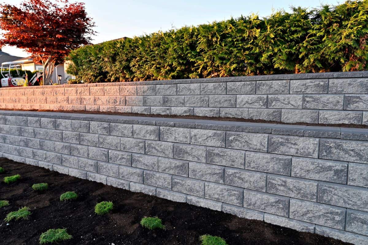 A close-up capture of a gray colored block stone retaining wall built as a two tier wall into an existing garden landscape viewed from the side in the early morning hours under sunny conditions.