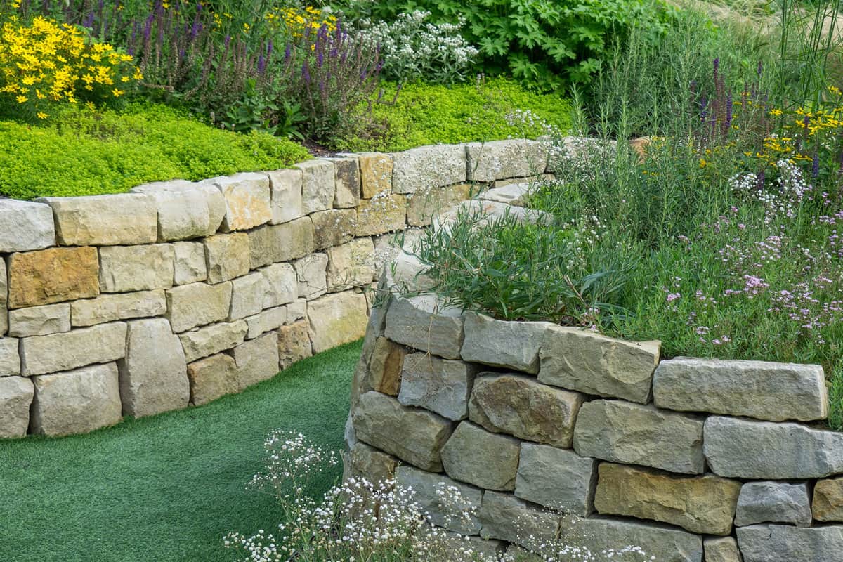 A gorgeous retaining wall using square marble rock and flowers on top for landscaping purposes, Does A Retaining Wall Stop Water? [And How To Add Drainage]