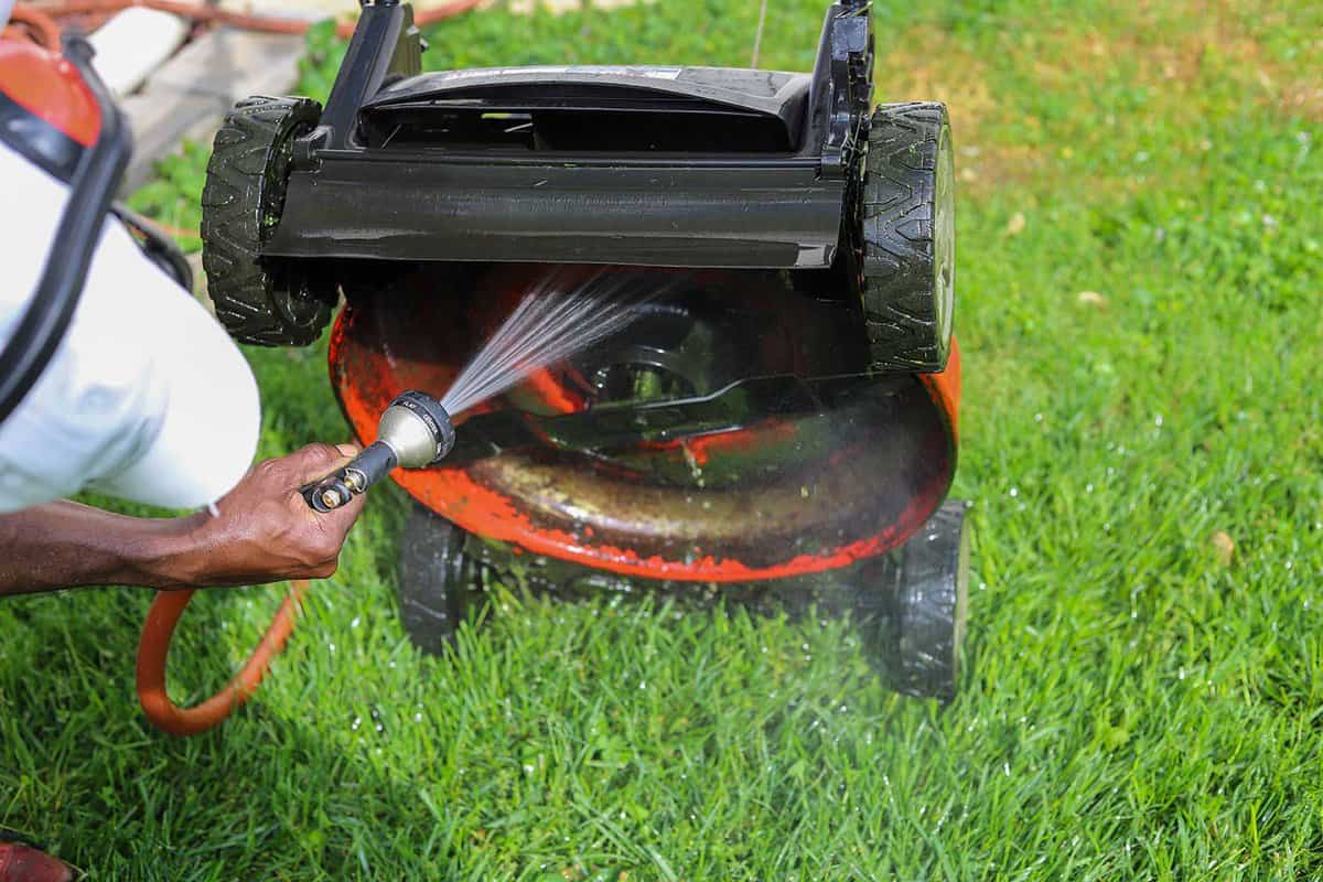 A man spraying and cleaning off a lawnmower with a water hose outside in the summer