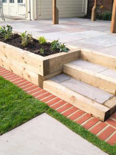New steps in a garden or back yard leading to a raised patio, alongside a new raised flowerbed made using wooden sleepers. A mowing strip of bricks is in front of newly laid turf. - How To Edge A Raised Patio [15 Gorgeous Ideas!]