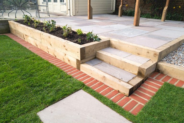 New steps in a garden or back yard leading to a raised patio, alongside a new raised flowerbed made using wooden sleepers. A mowing strip of bricks is in front of newly laid turf. - How To Edge A Raised Patio [15 Gorgeous Ideas!]