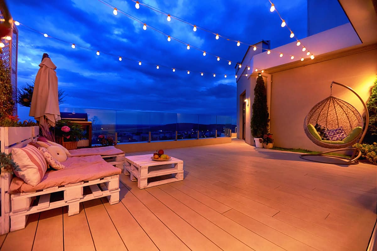 cozy rooftop patio area with lounge zone, hanging chair and and string lights at warm summer evening