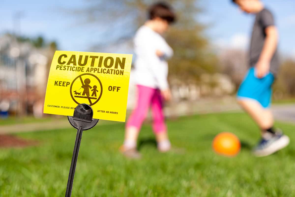 A yellow yard sign warning kids and pets of the recent pesticide spraying and advices them to stay away.