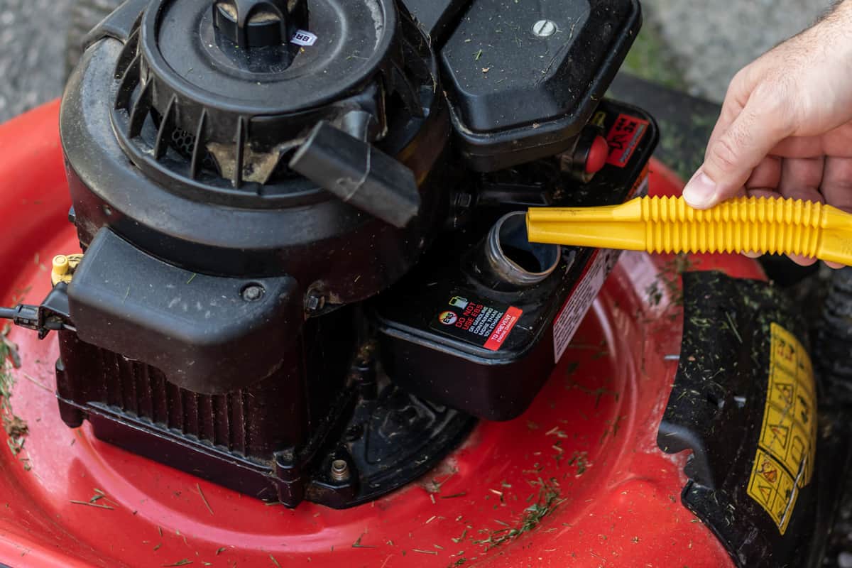 Closeup of man filling up red lawnmower with gasoline from a can with a yellow spout. Lawn and grass clippings on the mower.