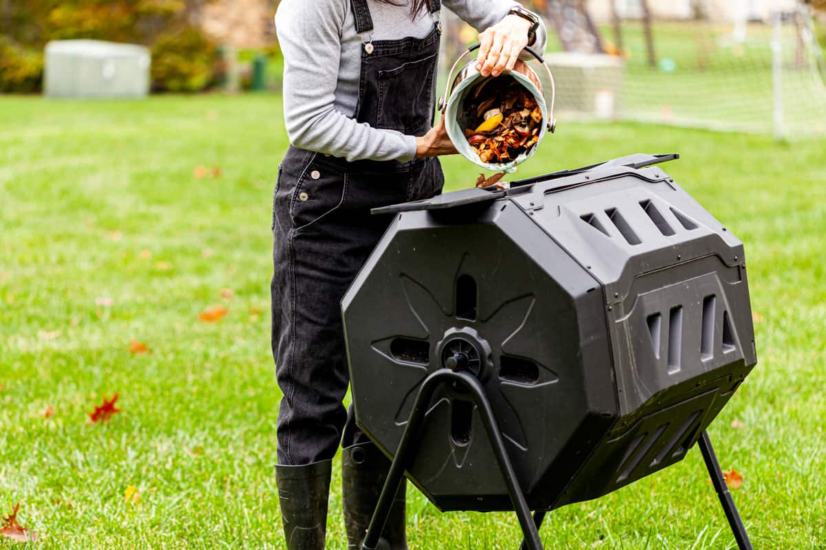 A woman is dumping a small bin of kitchen scraps into an outdoor tumbling composter