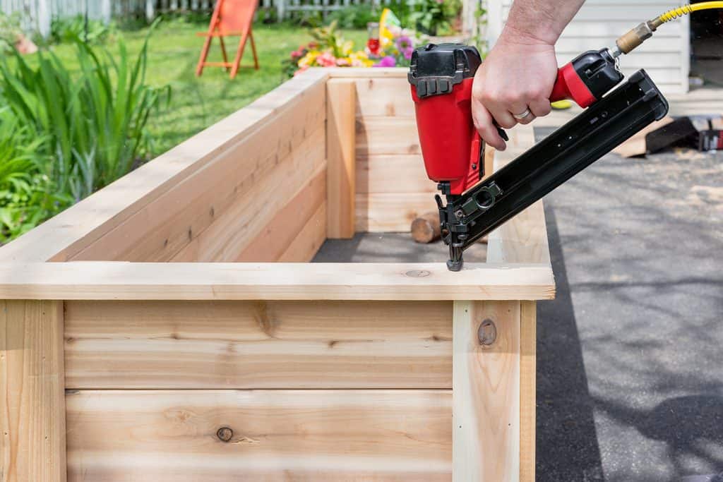 Close up of a man using a pneumatic nail gun to finish the trim on cedar garden planters with sawdust flying