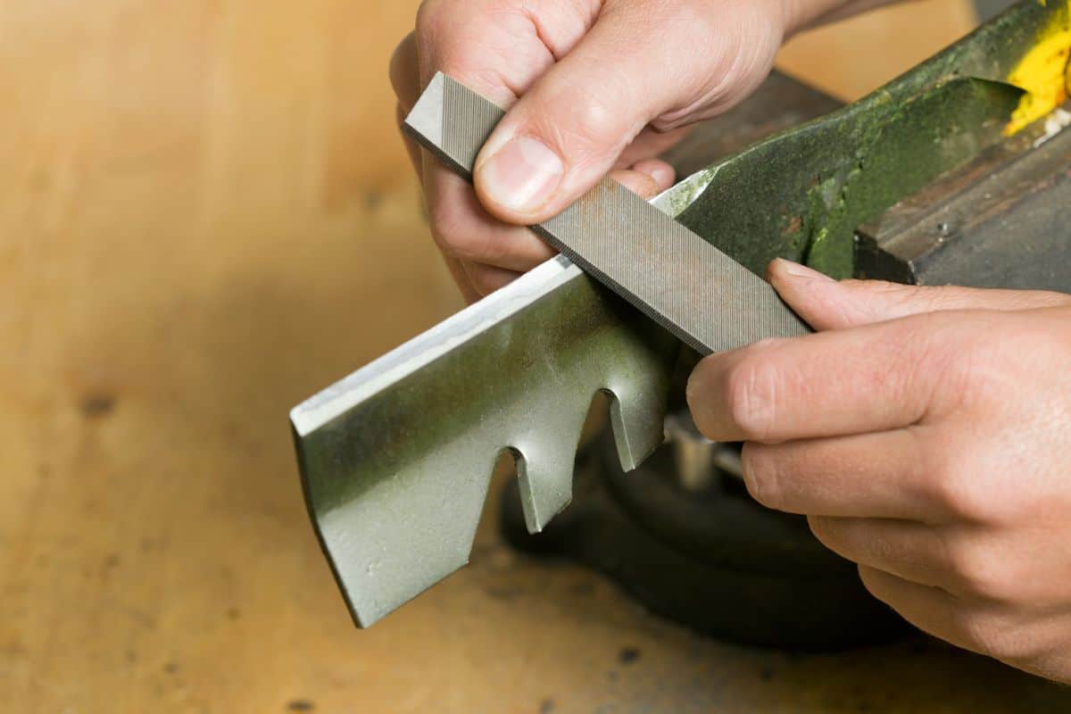 "Male hands are using a file to sharpen a lawnmower blade which is clamped in a vise atop a wood workbench. Focus is on the file near the bottom hand, the blade is slightly soft."
