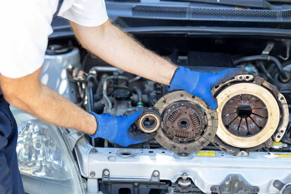 Automotive technician holding used car pressure plate, clutch disc and release bearing in front of the vehicle engine 