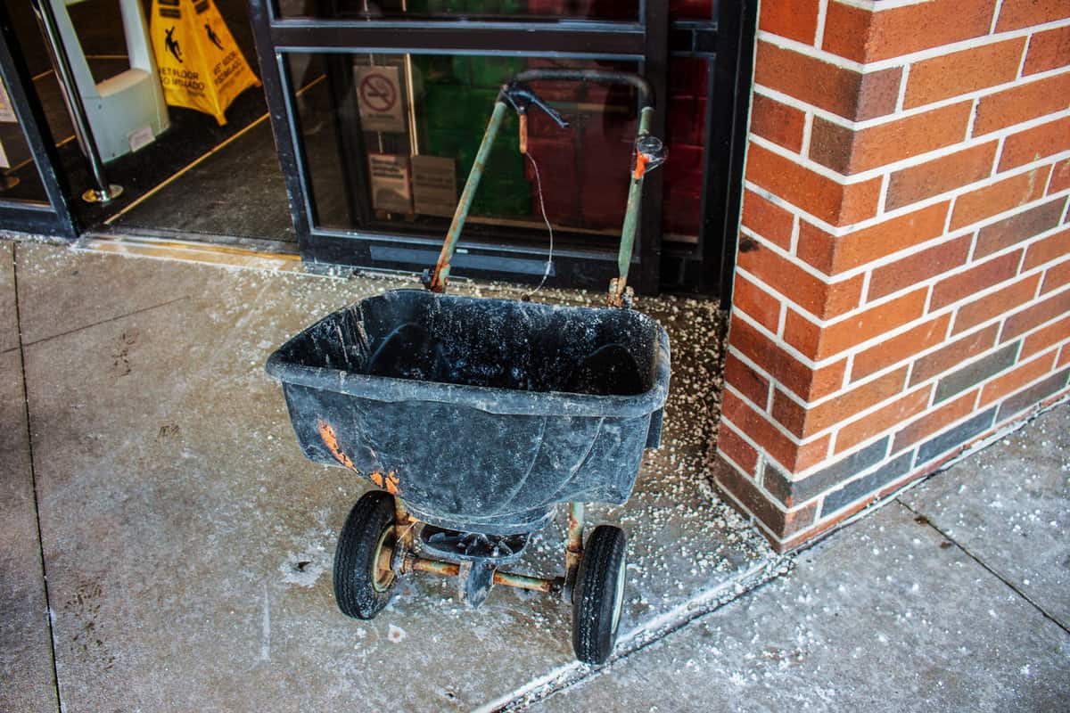 grungy two wheeled plastic salt spreader parked outside a store sliding glass door with salt scattered on the sidewalk around it after a snow 