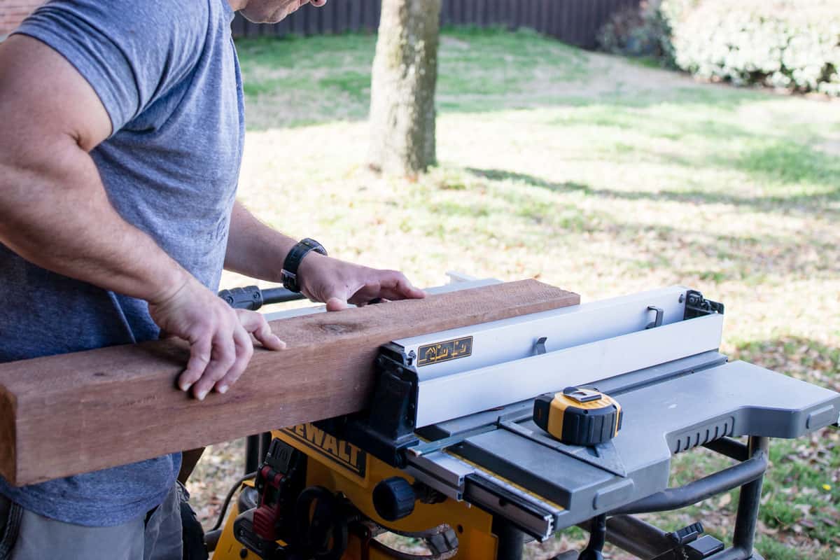 Side view of caucasian man in his yard guiding a 4x4 wooden post through the blade of a table saw that is cutting wood for a DIY proje