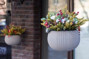 close up shot of a flower arrangement in a white basket pot hanging on blurred bokeh background. The flowers are yellow, red, pink with purple berries and white snow on them, How To Attach Hanging Baskets To Concrete Posts