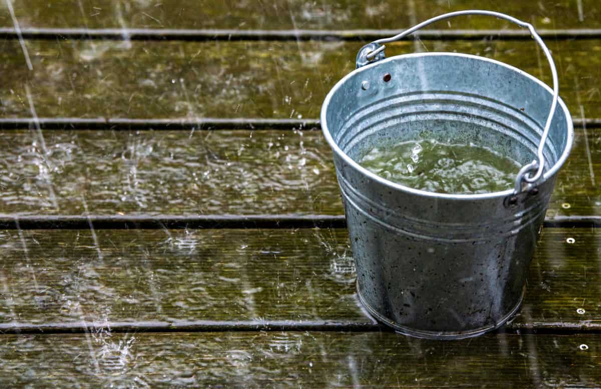 A silver aluminium bucket is standing on a green wood panels and heavy rain is visible