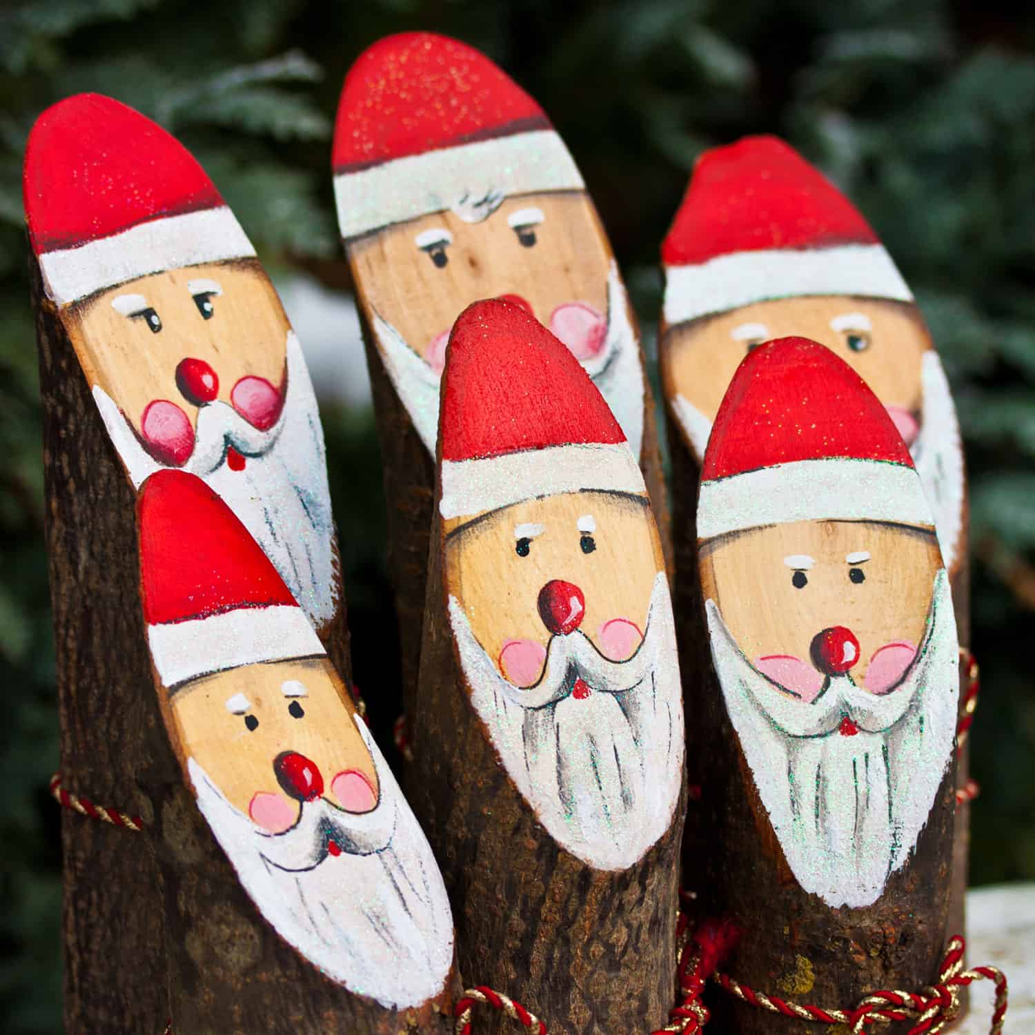 a trio of handcrafted Santa Claus figures carved into a vertically standing log, each sporting a red hat and a white beard with the largest Santa adorned with a green and red plaid ribbon scarf