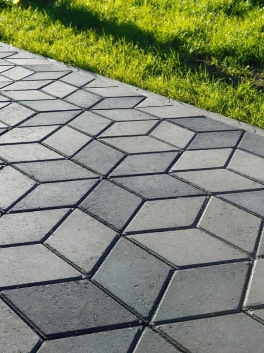 A close-up of a dark grey geometric concrete paver pathway, the angular shapes creating a pleasing pattern beside the vibrant green grass, illustrating a contrast between the meticulous hardscape and the soft, natural lawn ar 3:4