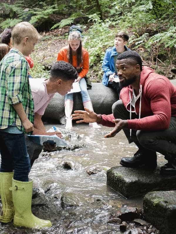 A diverse group of young students and an instructor engage in an outdoor educational activity by a stream, with the instructor gesturing expressively to explain a concept ar 3:4