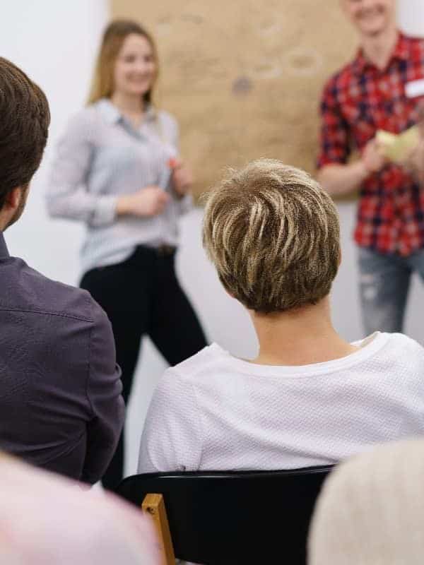 A gardening seminar where a woman in the background, smiling and gesturing, appears to lead an interactive session for an engaged audience whose backs are facing the camera ar 3:4