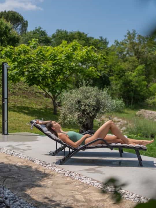 A person relaxes on a sun lounger, enjoying a tan in the sun on a concrete patio, surrounded by a lush garden with mature trees and shrubs, embodying a scene of peaceful outdoor leisure ar 3:4