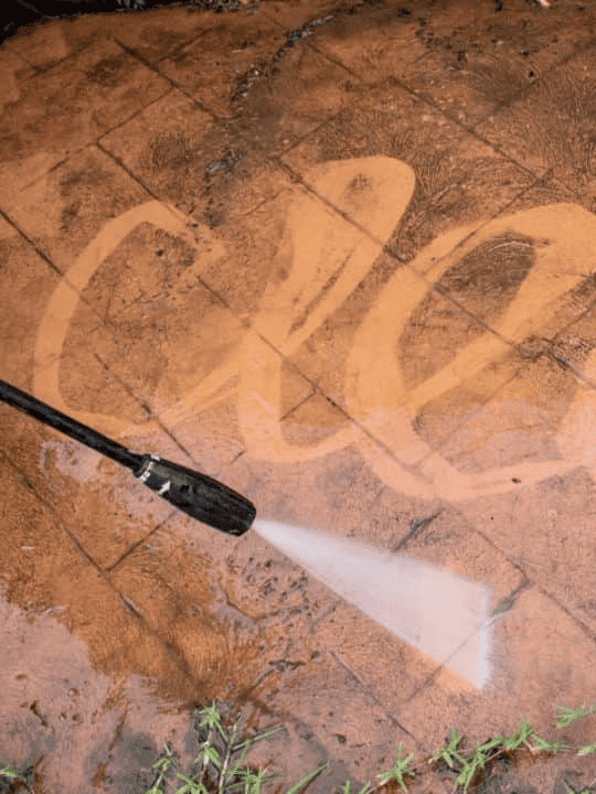 A pressure washer is in the process of cleaning a concrete patio, where the water's force is removing the surface dirt, revealing the unstained concrete beneath in a swoosh pattern, effectively demonstrating how staining does the fixing ar 3:4