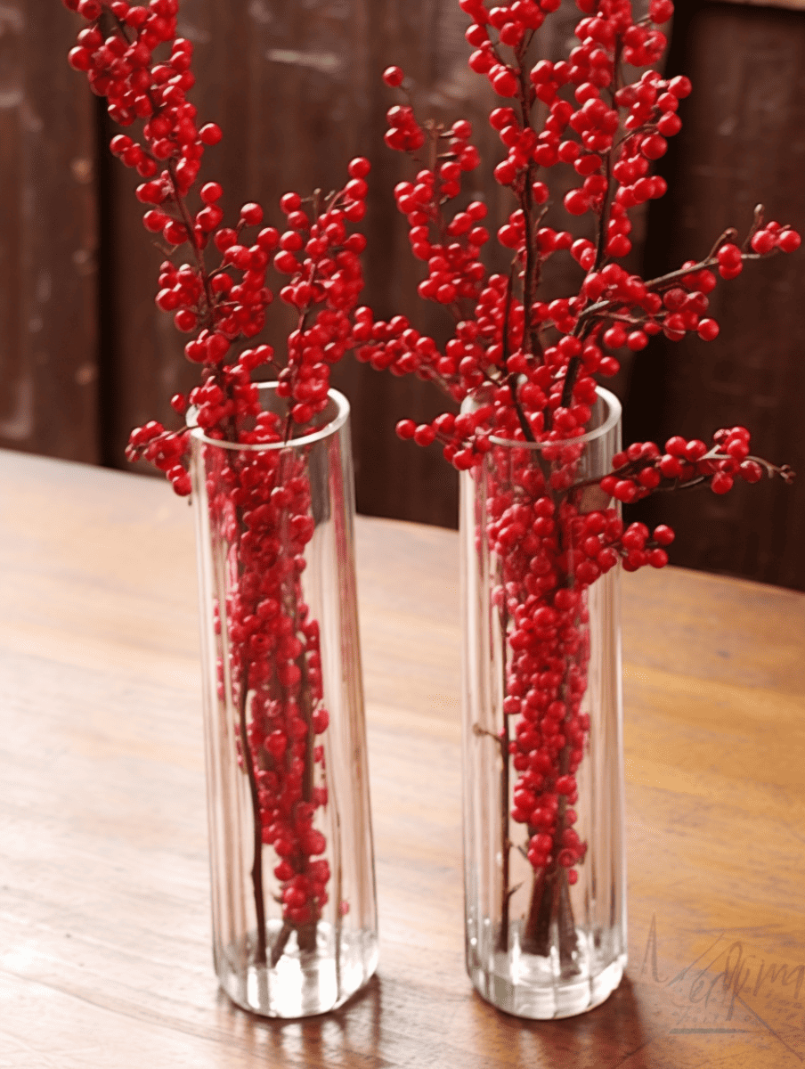 Two tall, slender, clear glass vases sit on a wooden surface, each filled with clusters of vibrant, small winter berries on branching stems, creating a striking contrast against the brown wood grain background