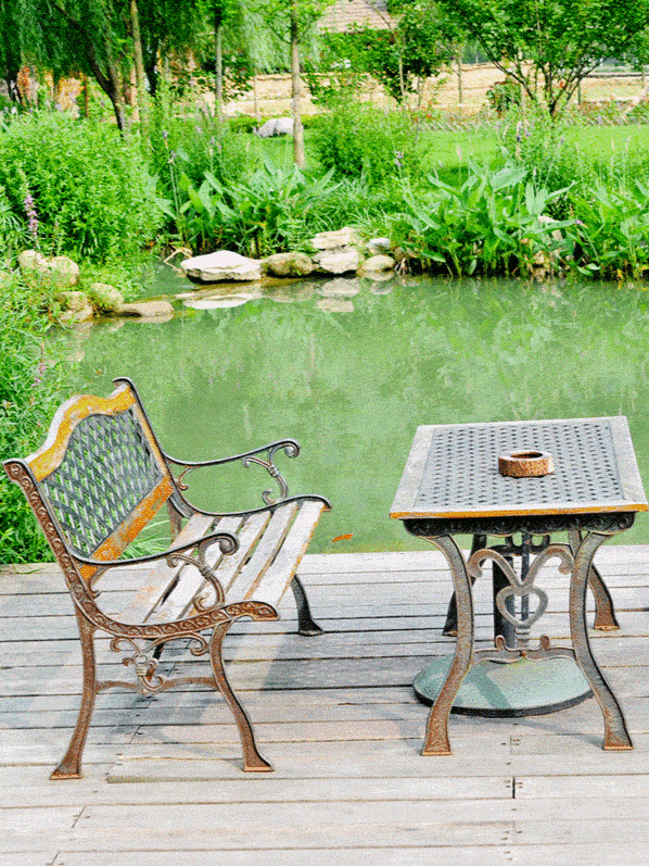 An ornate metal garden bench and matching table sit on a wooden platform by a serene pond, amidst a lush green landscape, inviting quiet reflection or a peaceful outdoor break ar 3:4