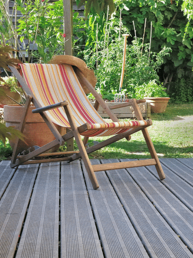 A wooden deck chair with colorful stripes is positioned invitingly on a grey deck, with rustic terracotta pots and garden trellises supporting climbing plants in the background, suggesting a casual garden setting ar 3:4