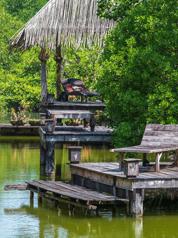 A rustic wooden dock with a thatched-roof gazebo and lounging chairs extends over tranquil waters, flanked by dense greenery, offering a serene spot for relaxation and contemplation ar 3:4